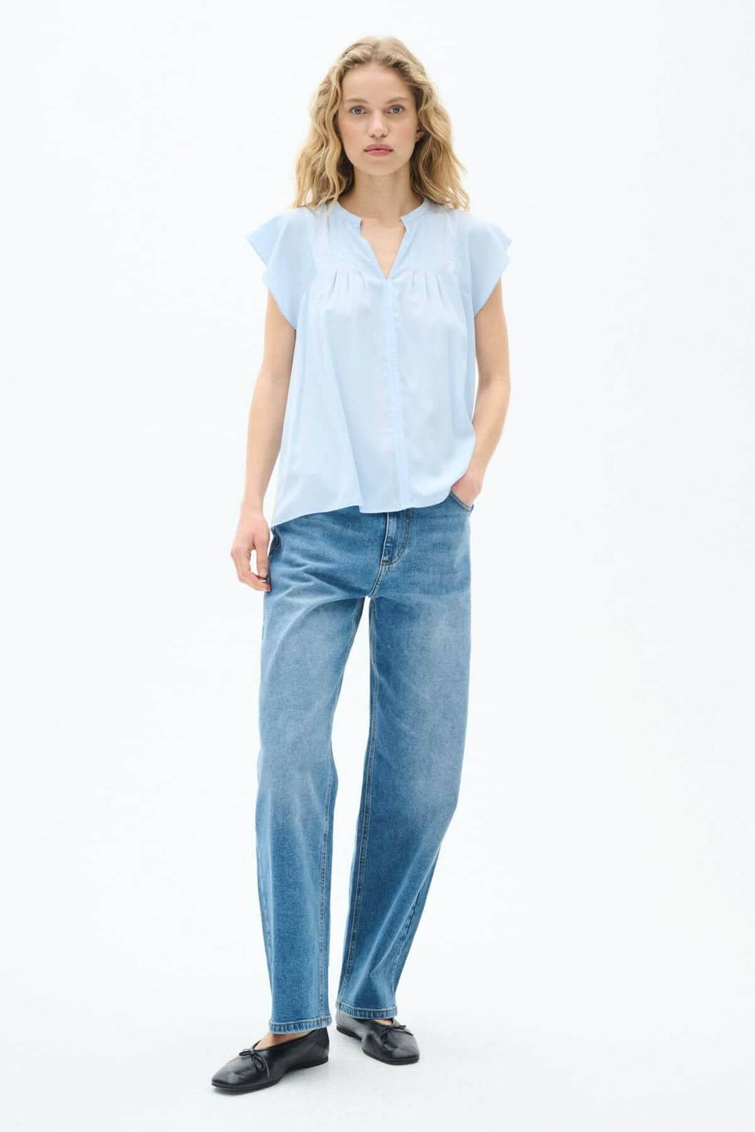 Modèle en blouse légère bleue, manches volantes et coupe fluide, portée avec un jean et des chaussures noires.