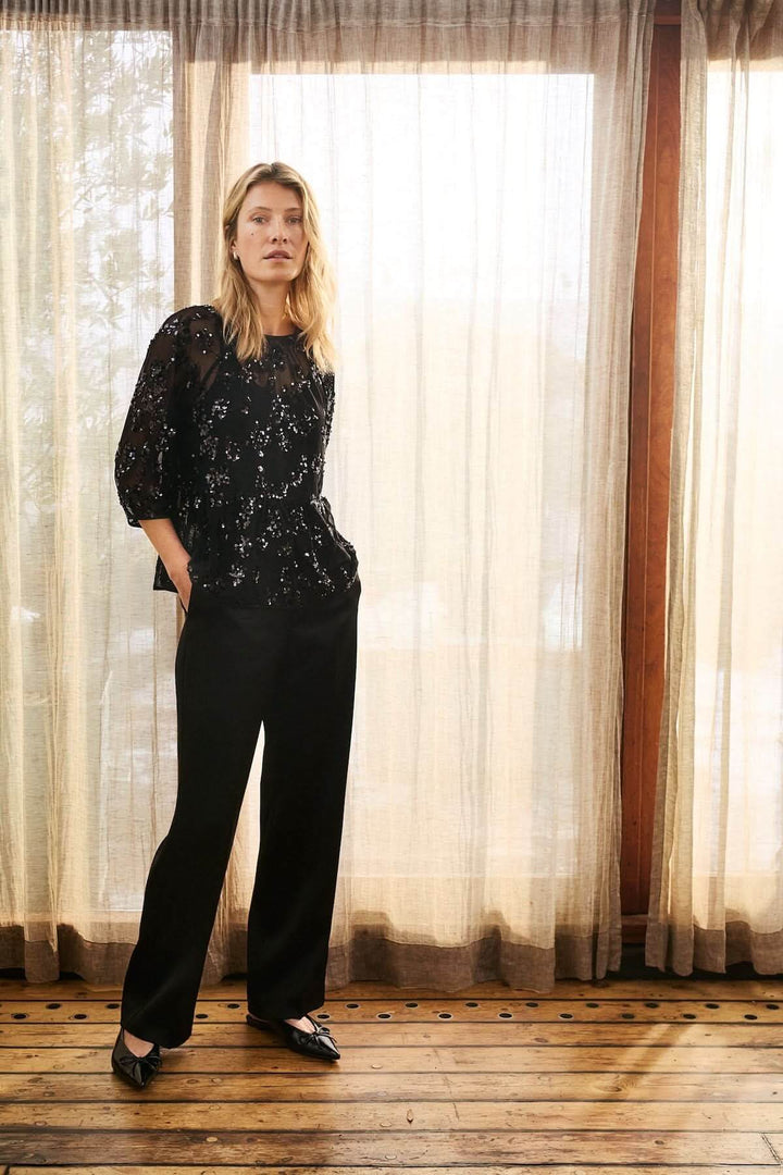 Blouse Vivan à sequins floraux portée avec pantalon noir, élégance raffinée sur fond de rideaux en lumière naturelle.