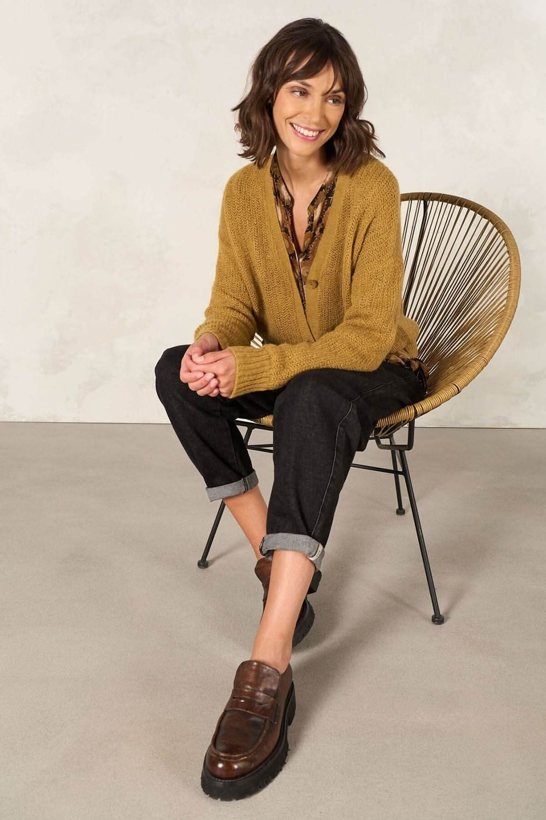 Femme souriante portant un cardigan moutarde et pantalon noir, assise sur une chaise en rotin.