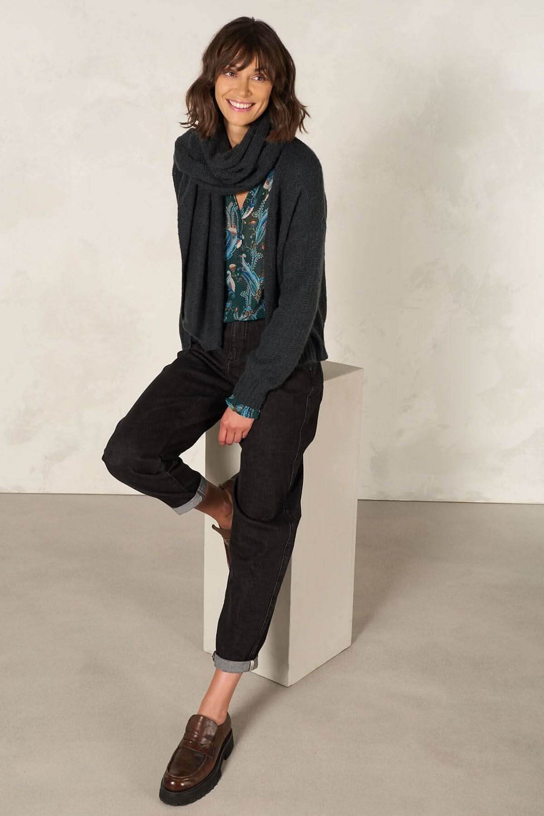Femme souriante portant un pull noir, un jean noir et un foulard, posant sur un support en béton.