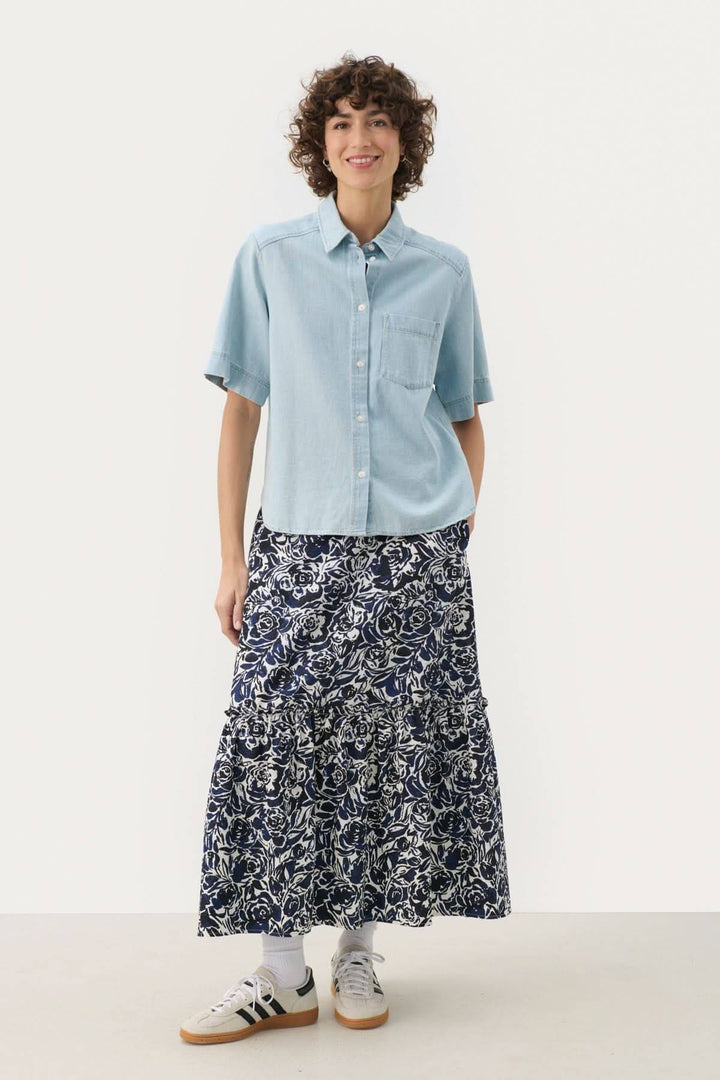 Femme portant la blouse en jeans Nicla, associée à une jupe longue et des baskets pour un look décontracté.