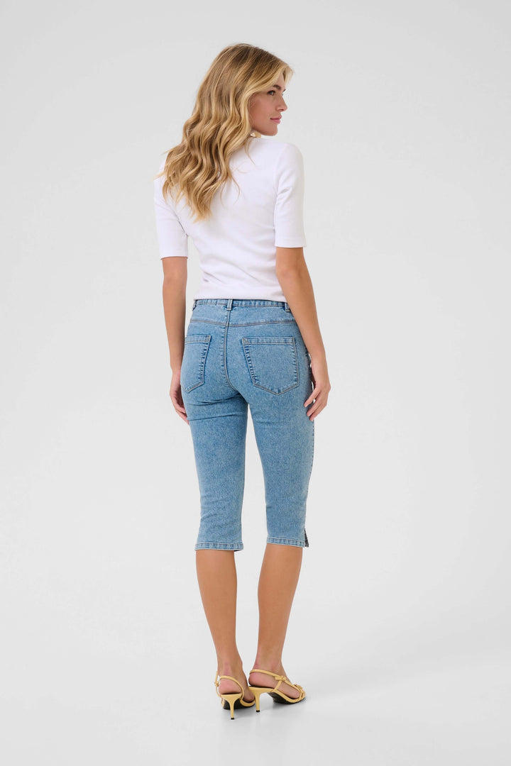 Femme en capri denim extensible ajusté, longueur sous le genou, vu de dos, portant un haut blanc et des sandales.
