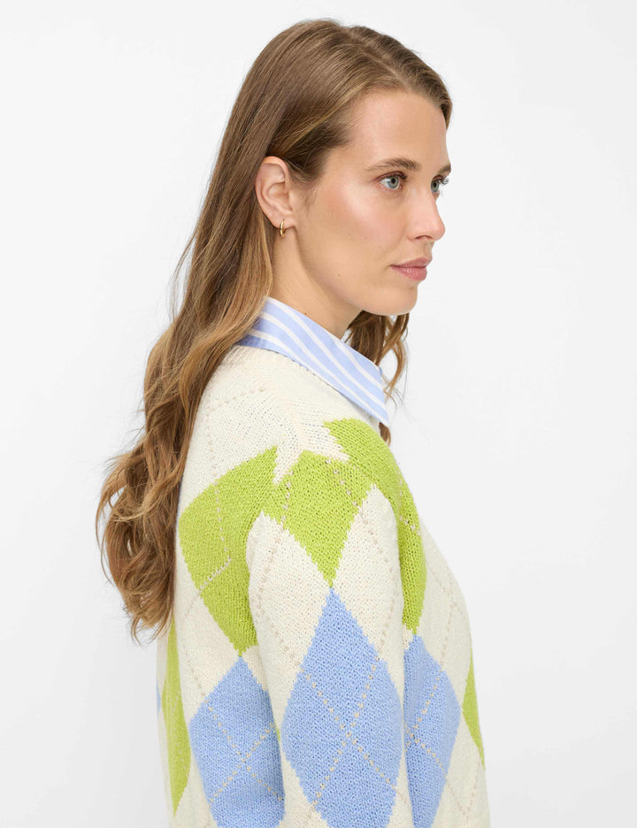 Femme portant un cardigan court à motif losanges ANN, tricoté avec des losanges verts et bleus, ajoutant une touche rétro et élégante.