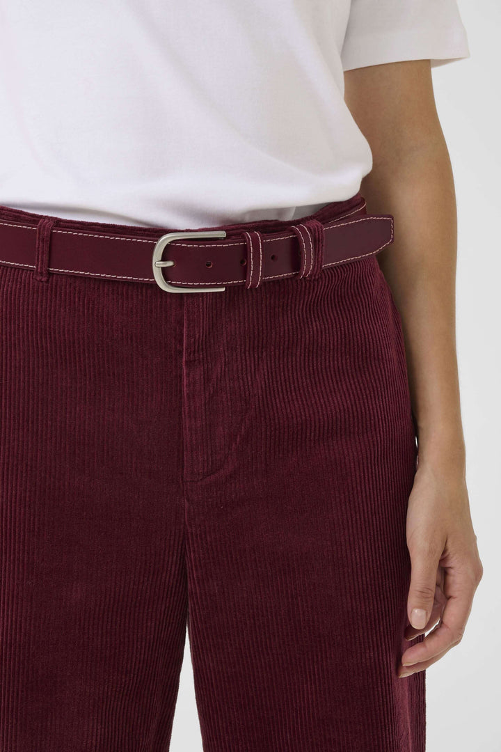 Ceinture Seyra en cuir véritable bordeaux avec surpiqûres contrastantes et boucle métallique, portée sur un pantalon en velours côtelé rouge.