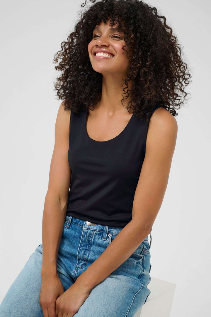 Femme souriante portant un débardeur noir et un jean, assise sur un tabouret blanc
