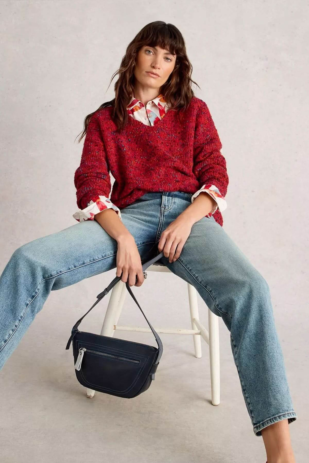 Femme portant un chandail col en V rouge avec une chemise à motif et un jean, assise sur une chaise avec un sac à main noir élégant.