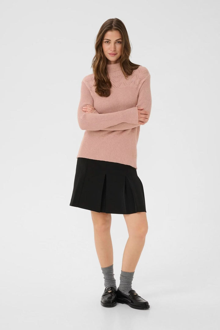 Femme portant un chandail à col montant rose avec une jupe noire, illustrant un style moderne et raffiné.