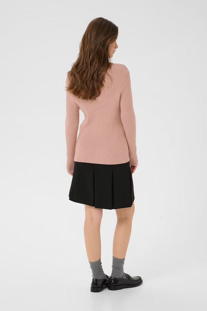 Femme portant un chandail à col montant côtelé en rose avec une jupe noire, vue de dos, style moderne et élégant.