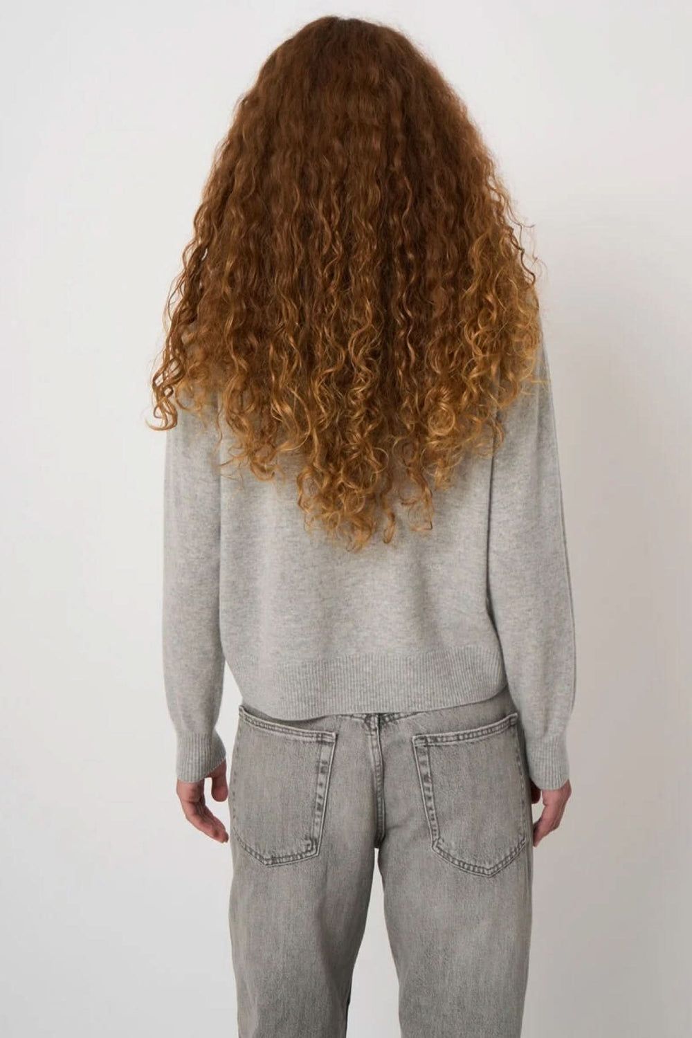 Vue arrière d'une femme portant un chandail en tricot gris avec motif géométrique, silhouette droite en jean décontracté, cheveux longs bouclés.