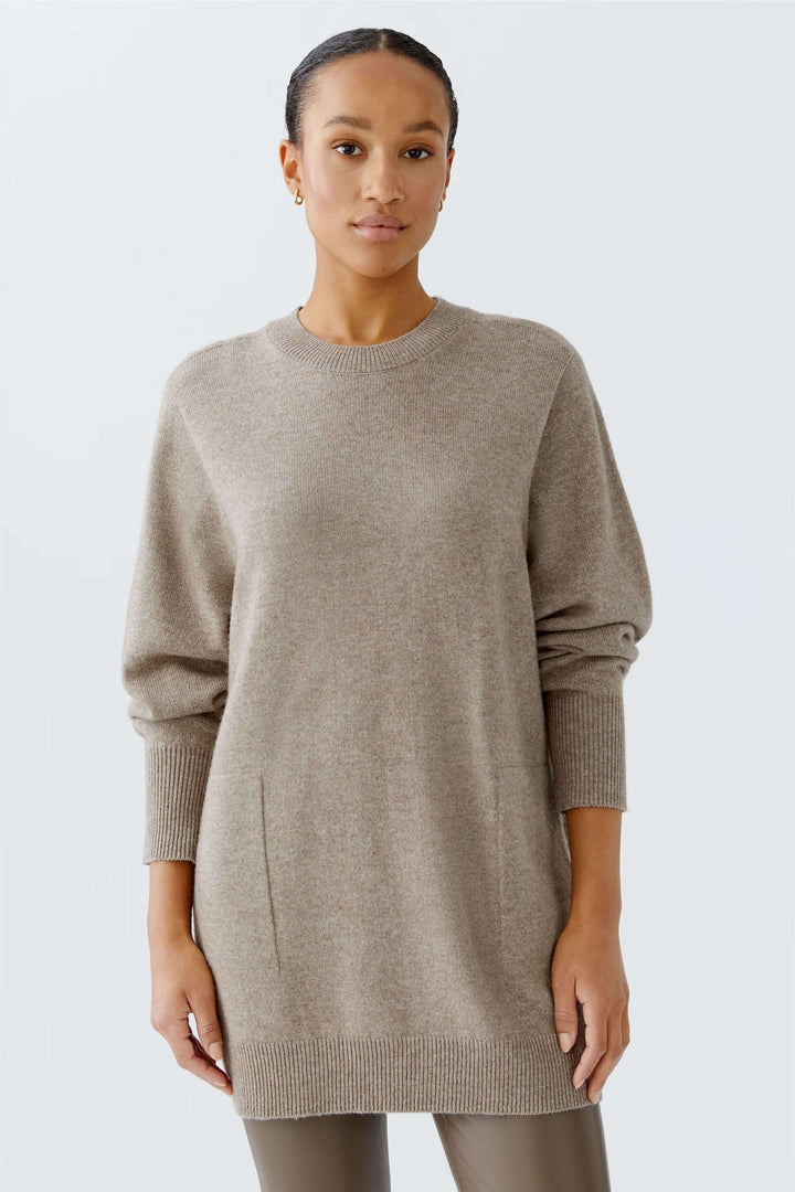 Femme portant un chandail long ample à col rond, couleur beige, manches longues, design minimaliste et confortable.