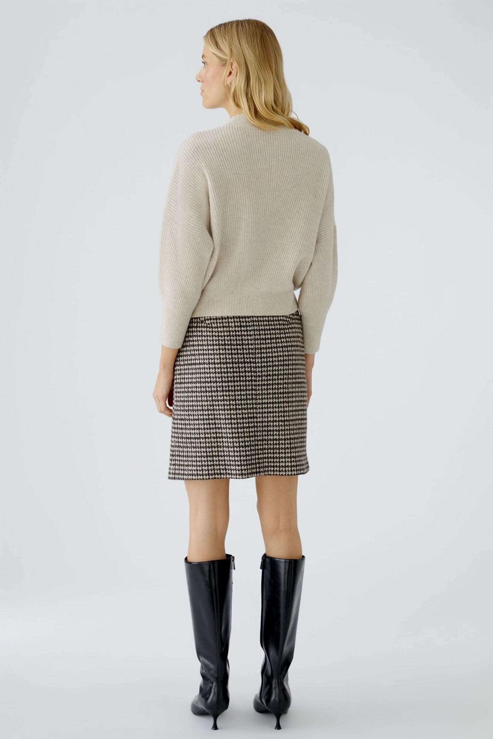 Femme portant un chandail maille fine avec jupe à motifs et bottes noires, vue de dos.