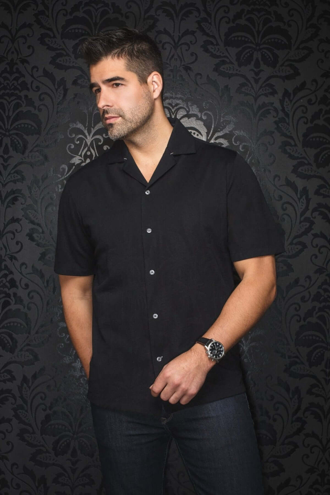 Homme portant une chemise noire à manches courtes avec un col cubain, sur fond décoratif élégant.