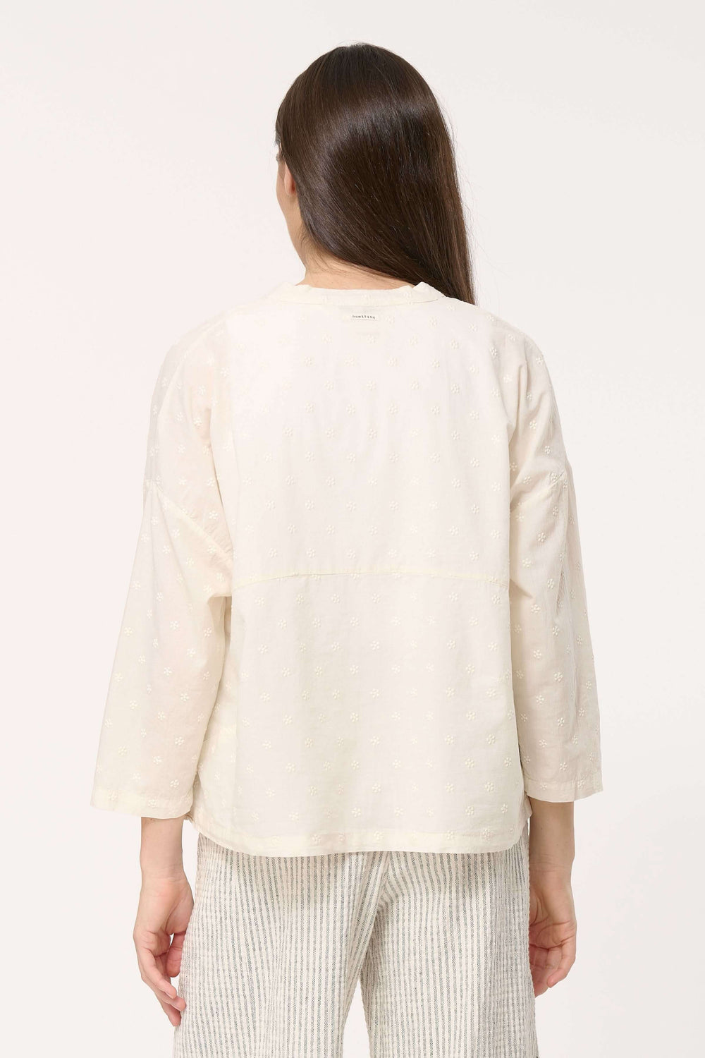 Femme portant une chemise légère en coton brodé, vue de dos, avec un pantalon rayé, élégante et décontractée.