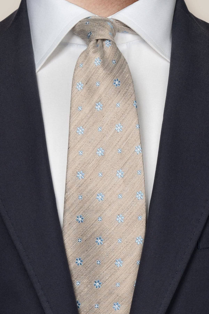 Cravate en soie et lin beige avec micro motifs floraux bleus, portée avec une chemise blanche et un costume noir.
