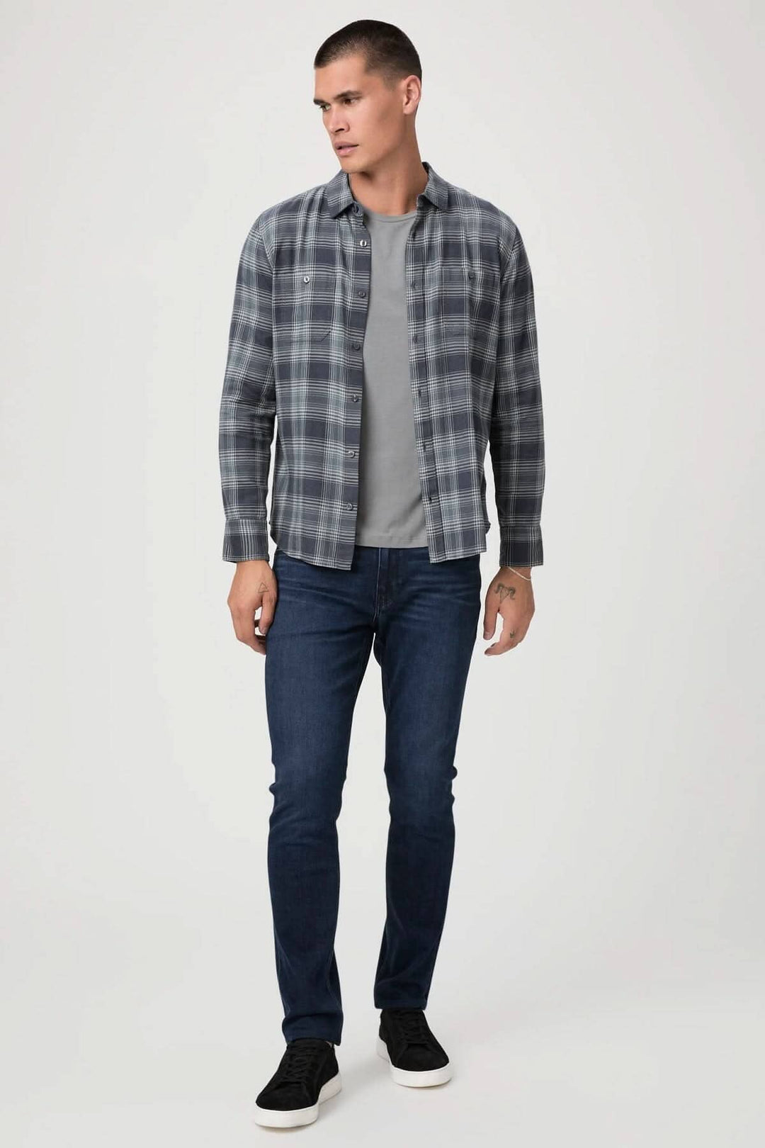Homme portant une chemise à carreaux grise et un t-shirt, associé à un jean ajusté bleu et des baskets noires.