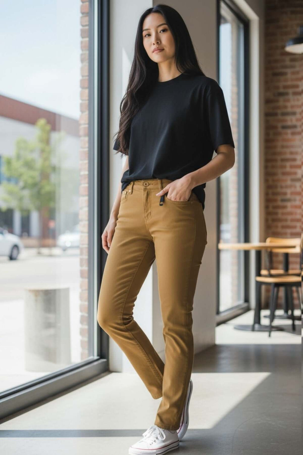 Femme porte un jean slim à taille mi-haute marron avec un t-shirt noir, debout près d'une fenêtre, style décontracté et moderne.
