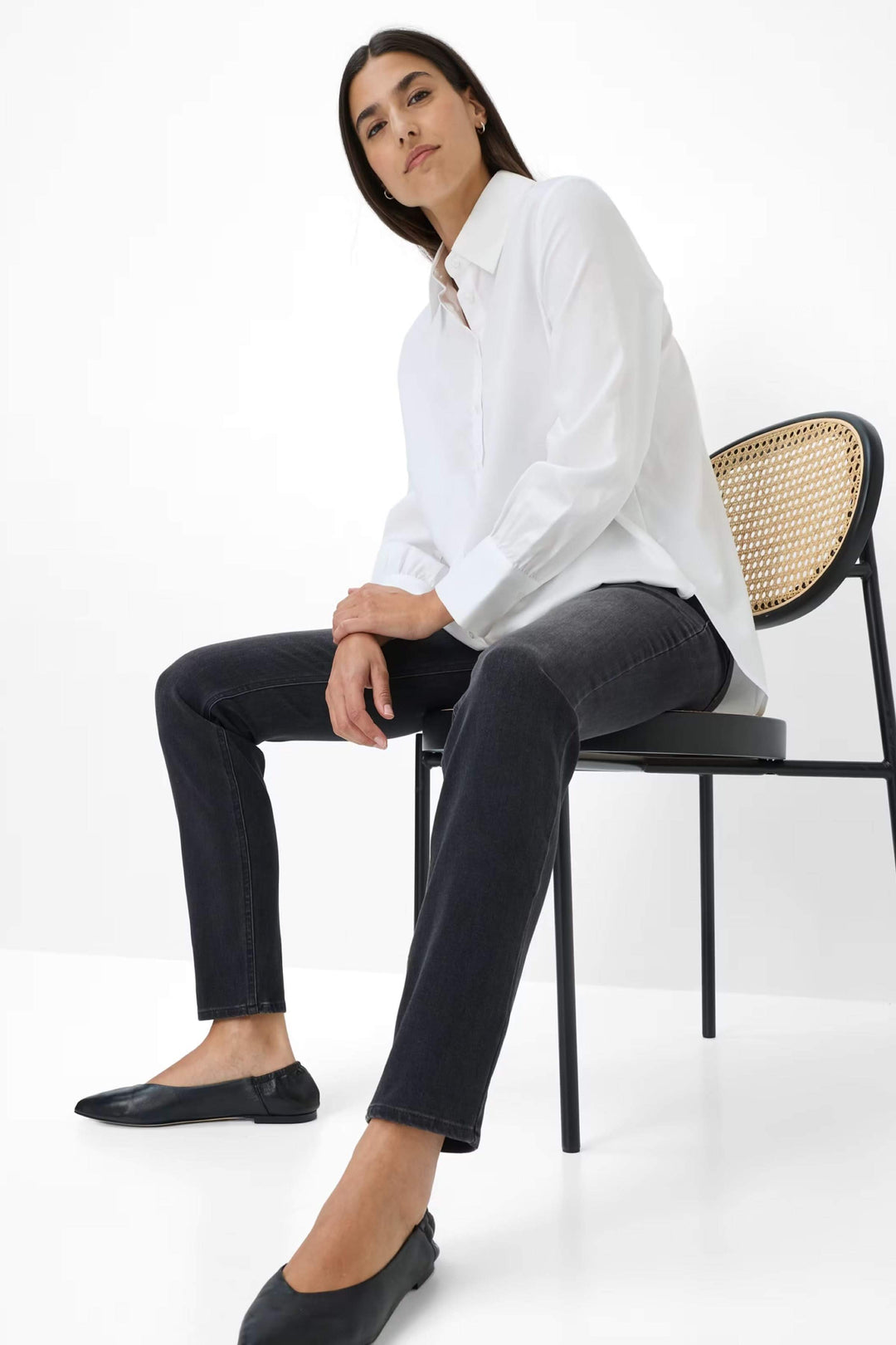 Femme portant Jeans Shakira coupe ajustée à taille mi-haute, assise sur une chaise, avec chemisier blanc et chaussures noires.