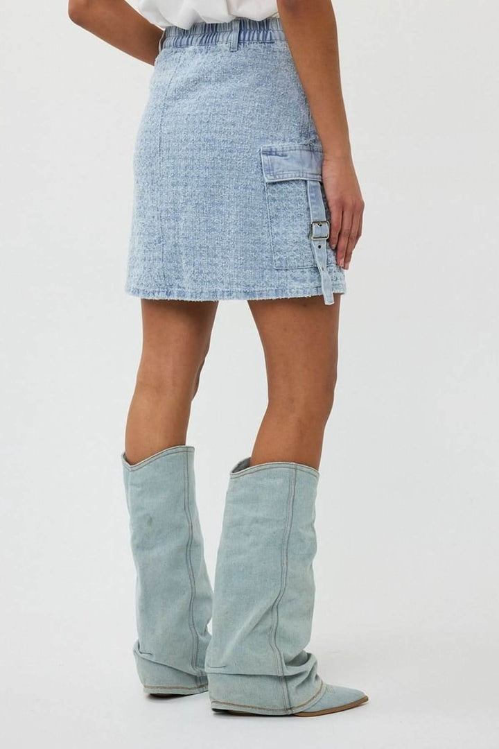 Jupe en denim texturé avec poche cargo latérale, coupe trapèze et sangle ajustable, portée avec des bottes.