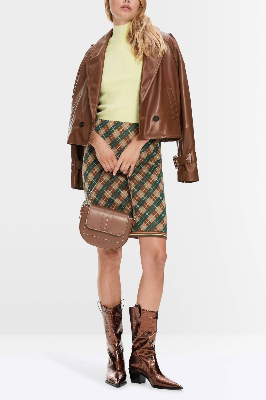 Modèle portant une mini-jupe à carreaux, un pull vert et une veste en cuir, avec des bottes brillantes.