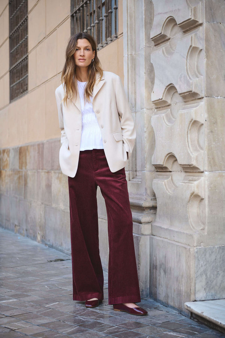 Femme portant le Pantalon Clarissi en velours côtelé avec coupe large et taille haute, style rétro-chic, sur fond de rue pavée.