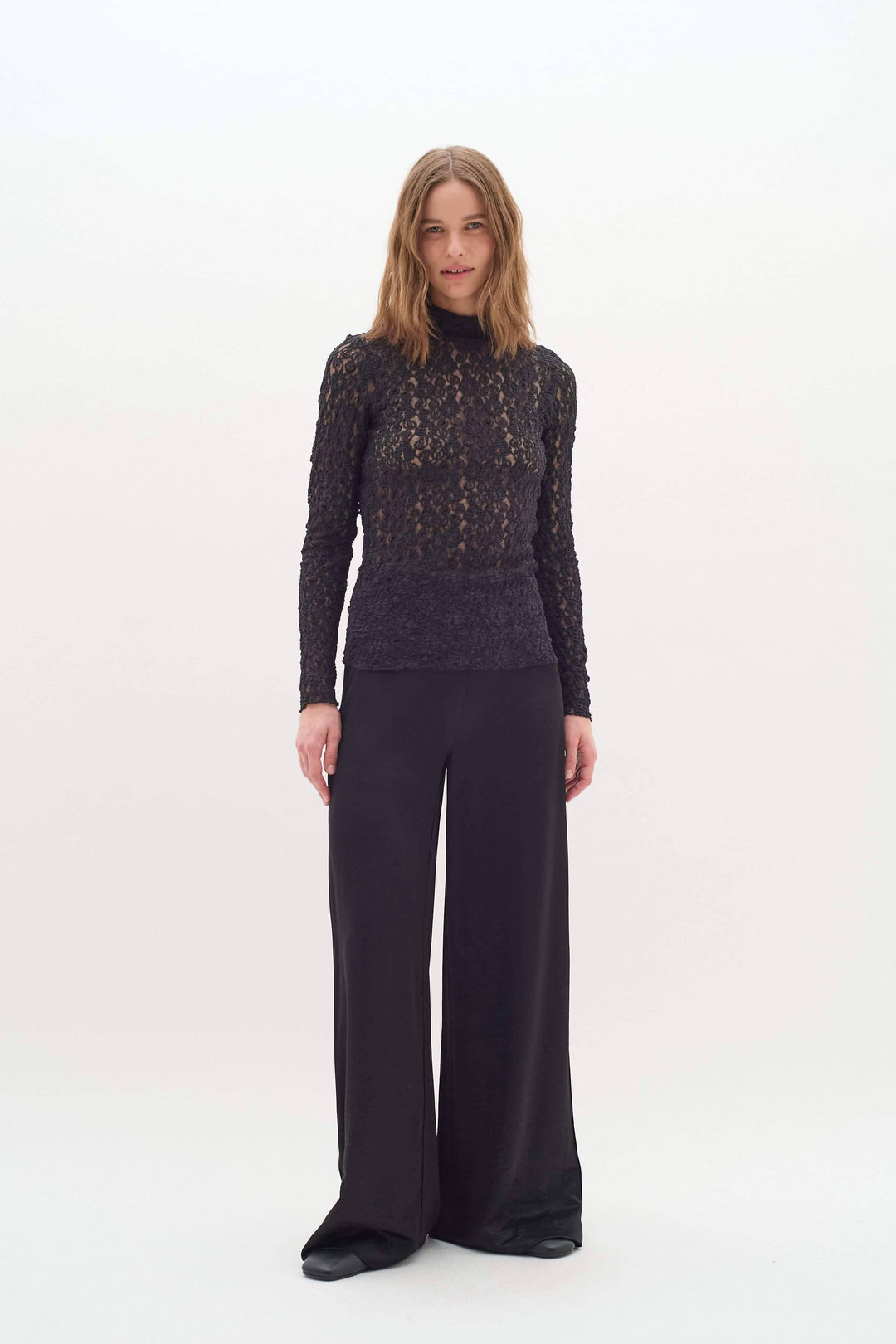 Femme portant un pantalon satiné noir et un haut en dentelle, style chic et moderne, sur fond blanc.
