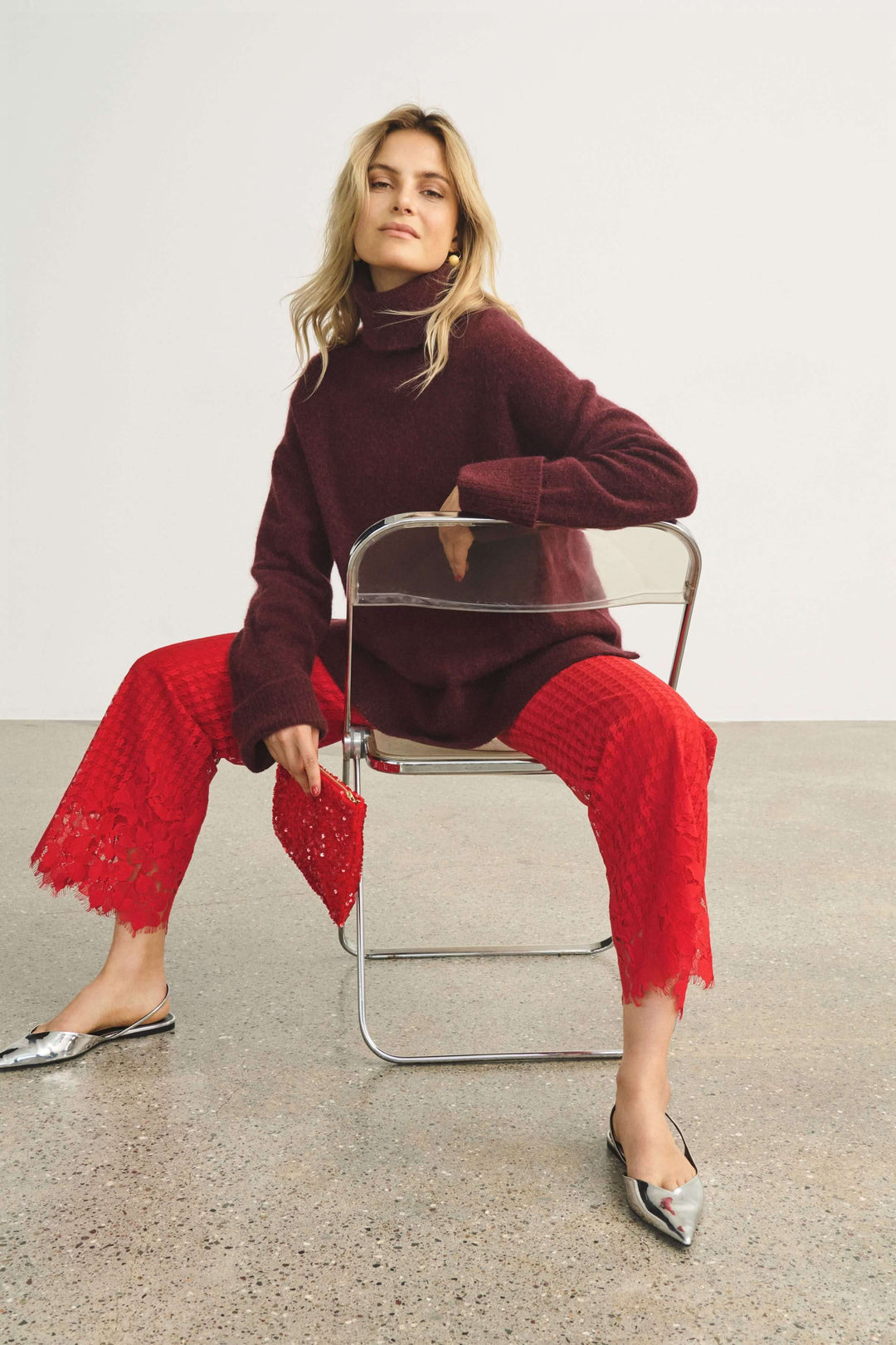 Femme élégante portant un pantalon en dentelle rouge et un pull à col roulé, assise sur une chaise.