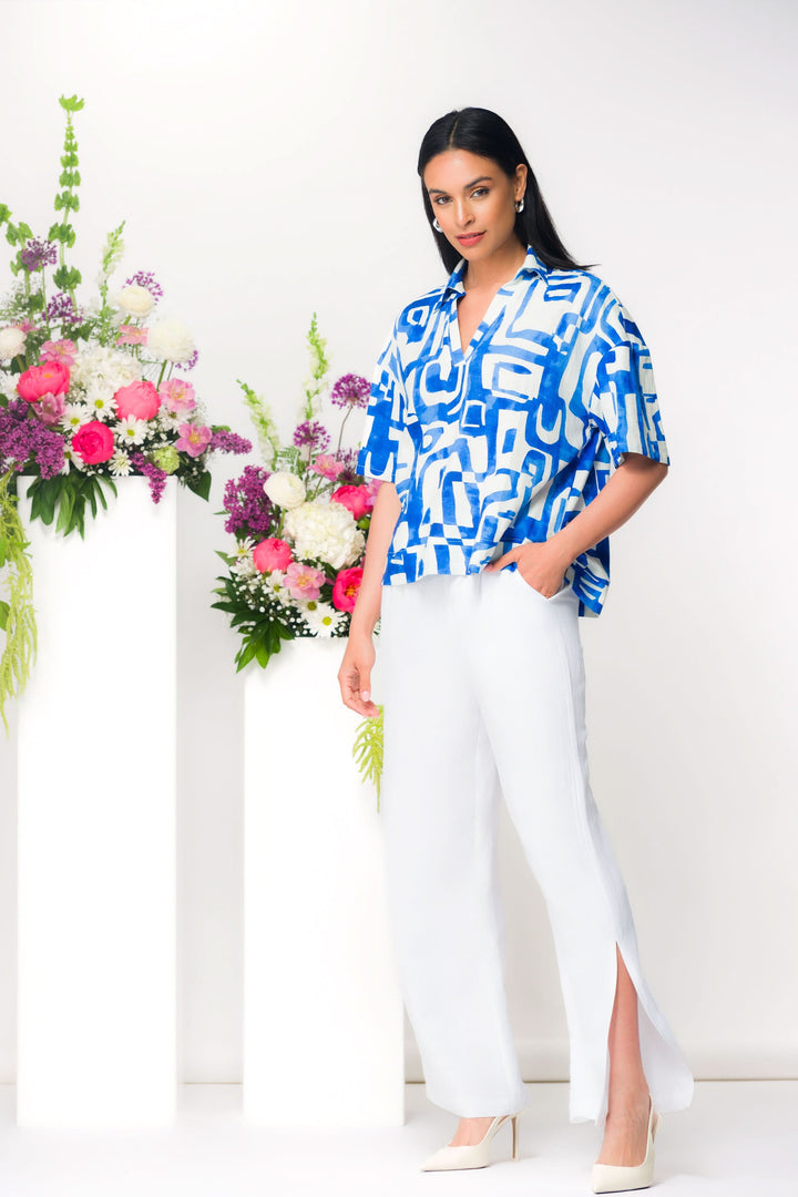 Femme portant un pantalon fluide à fente latérale blanc associé à un haut imprimé bleu et blanc, debout près de fleurs.