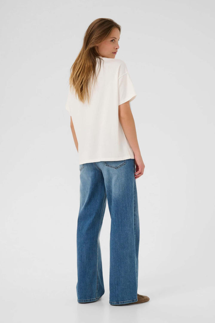Femme portant le T-shirt Helen blanc, à manches courtes et col rond classique, associé à un jean bleu pour un look décontracté.