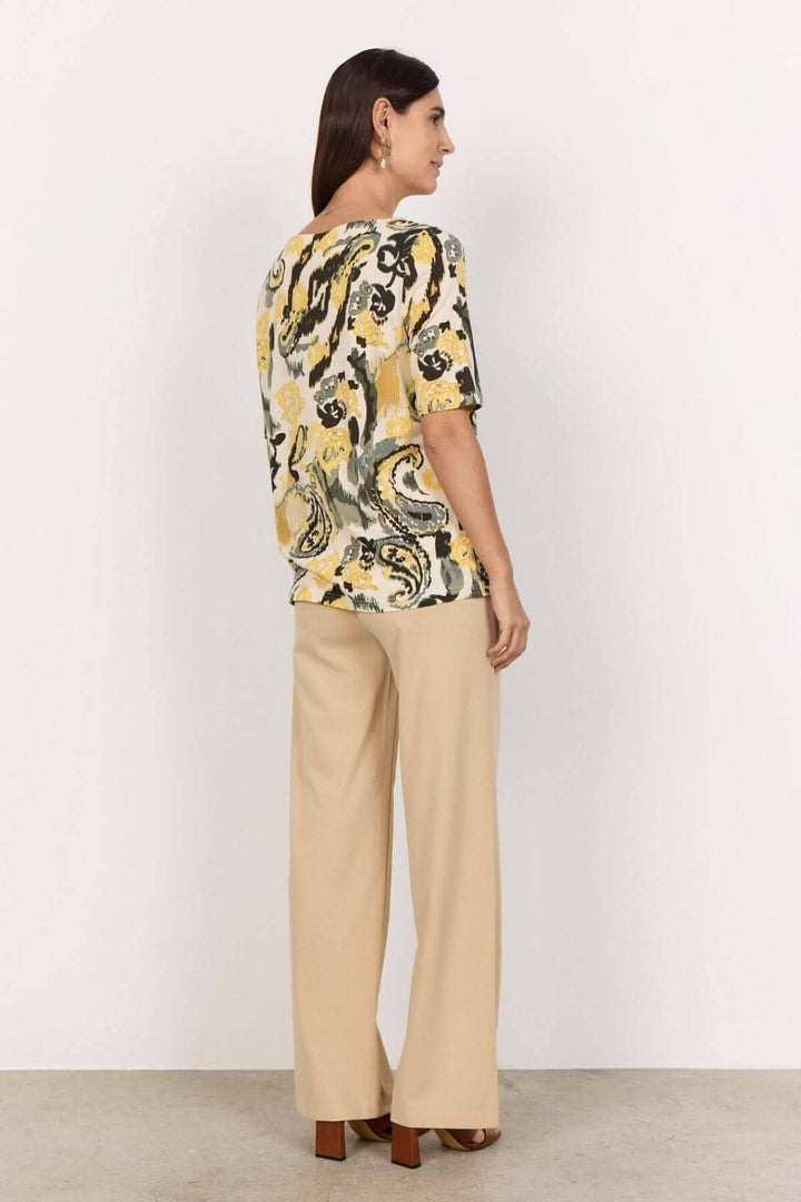 Femme vue de dos portant un T-shirt jaune imprimé abstrait avec un pantalon beige.