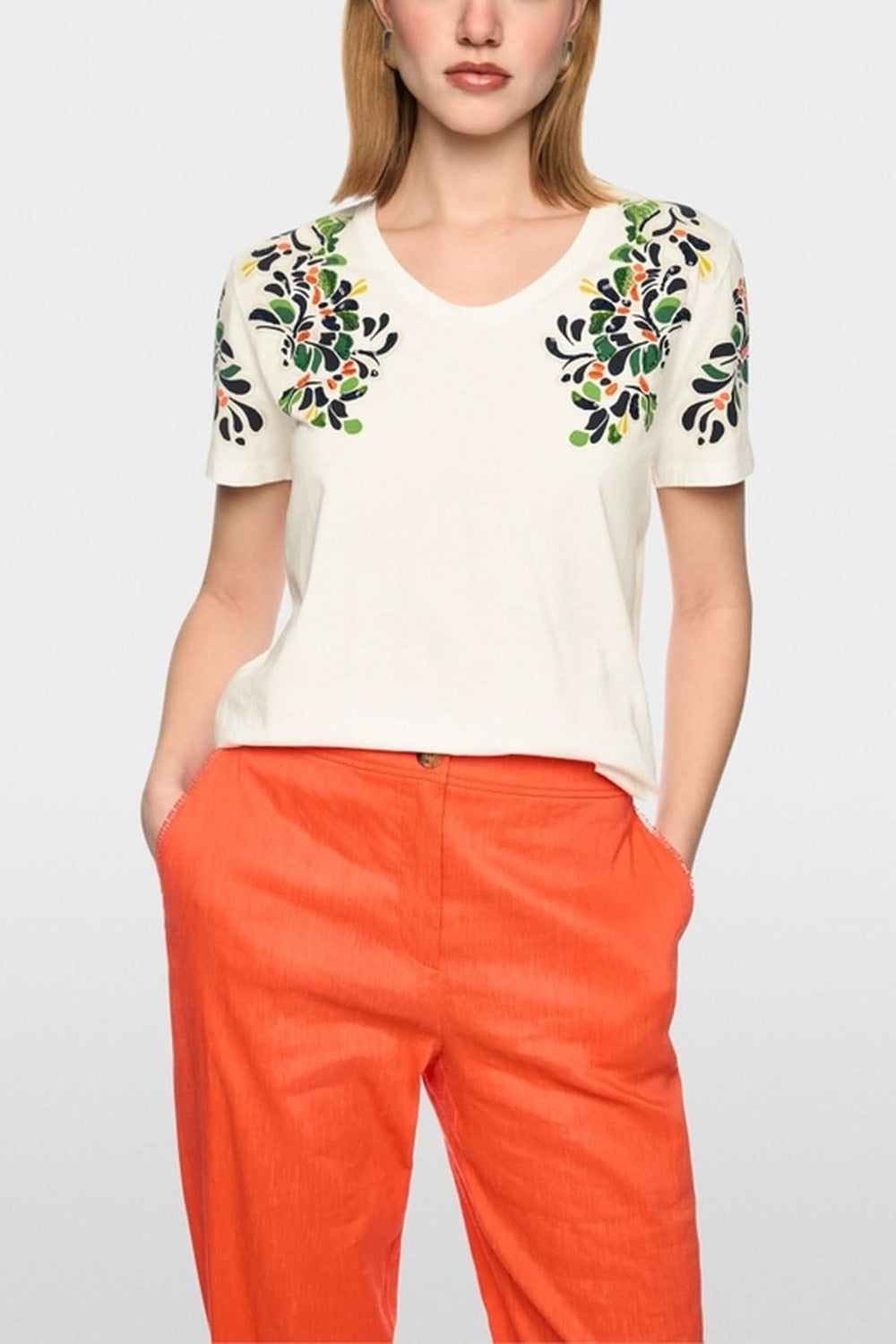 Femme portant un t-shirt imprimé hibiscus et paillettes, manches courtes et col rond, avec un design coloré, sur fond blanc.