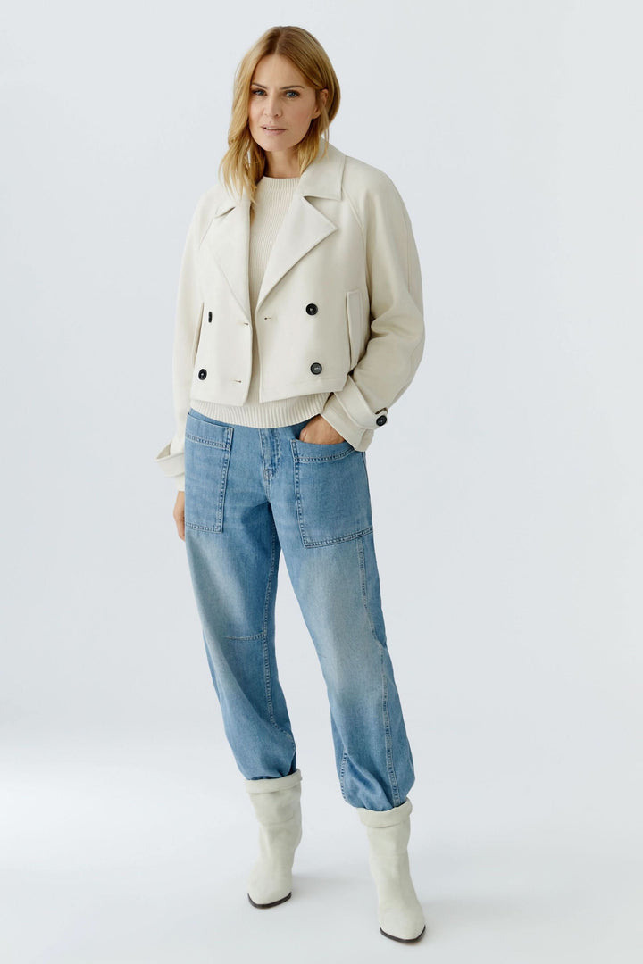 Veste courte croisée à col tailleur, style structuré et moderne, portée par une femme avec un jean décontracté et des bottes blanches.