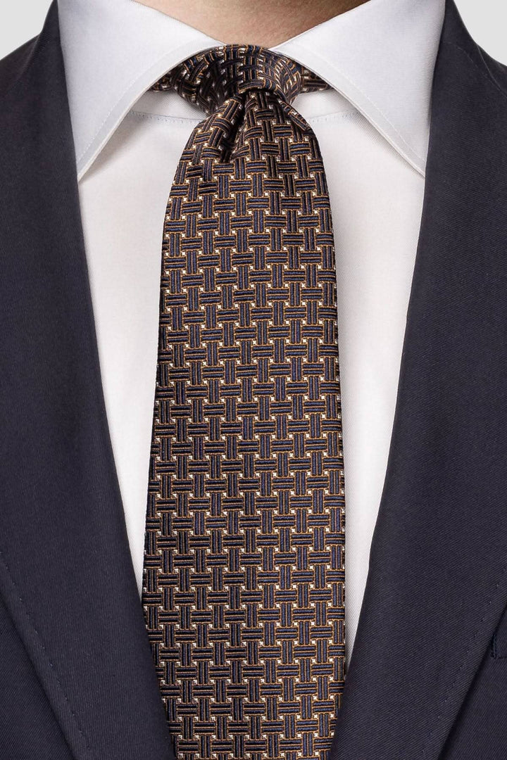 Cravate en soie avec motif tressé bleu marine, doré et blanc, portée avec une chemise blanche et un costume sombre.