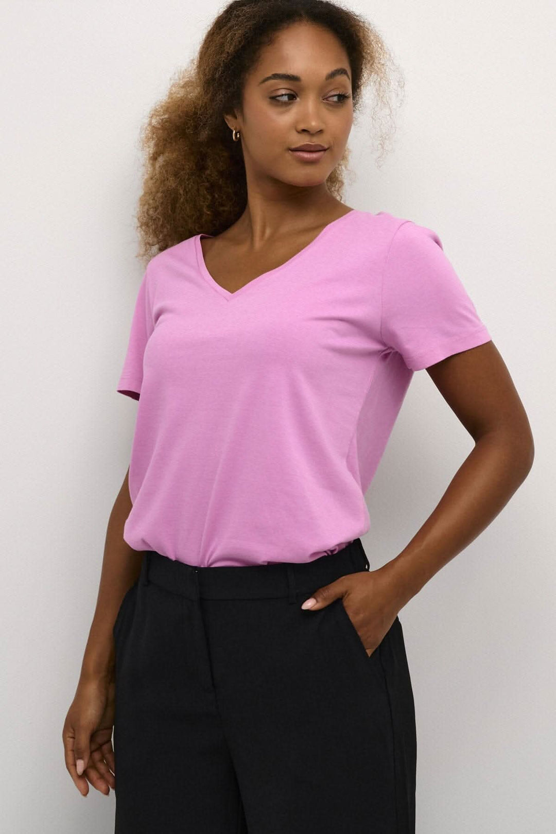 T-shirt Naia rose avec encolure en V et manches courtes, porté avec un pantalon noir.