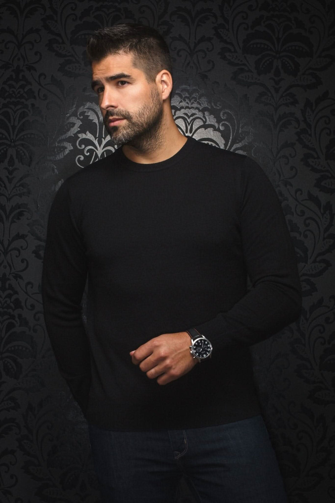 Homme portant un chandail col rond en mérinos noir, avec montre au poignet, sur fond orné.