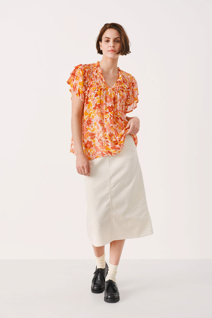 Blouse fleurie Sinna avec manches volantes, associée à une jupe blanche, sur fond neutre.