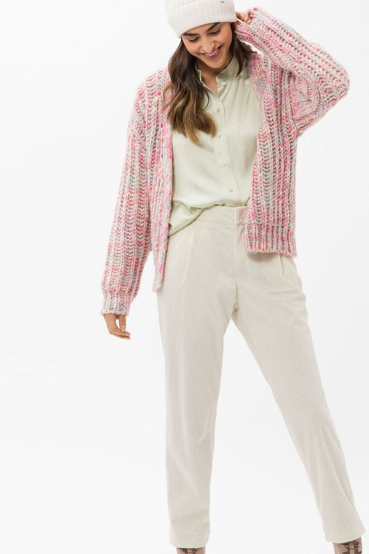 Femme portant un cardigan rose tricoté et un pantalon beige avec un chapeau. Style décontracté et confortable.
