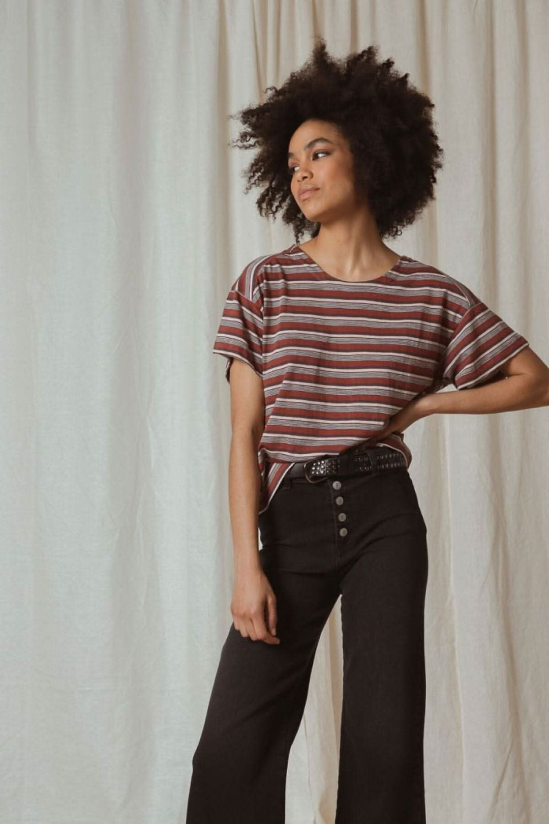 Femme avec des cheveux crépus portant un t-shirt à rayures et un pantalon noir, devant un fond clair.