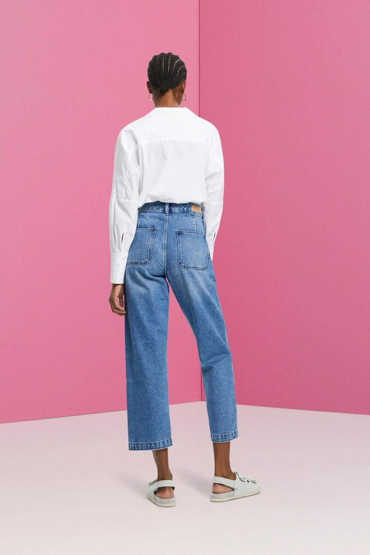 Femme portant un pantalon Dad en denim avec une chemise blanche, arrière, devant un fond rose.