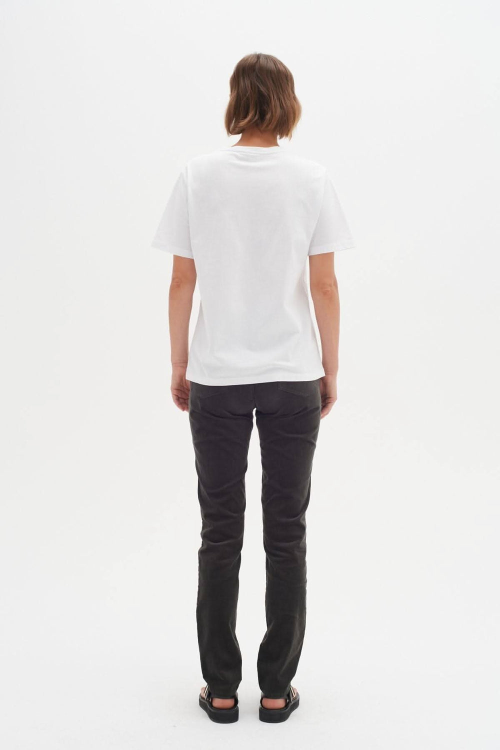 Femme vue de dos portant un t-shirt blanc et un jean foncé, sur fond blanc.