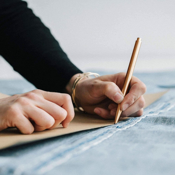 Person sketching on paper over denim fabric with a stylish gold pen in hand.