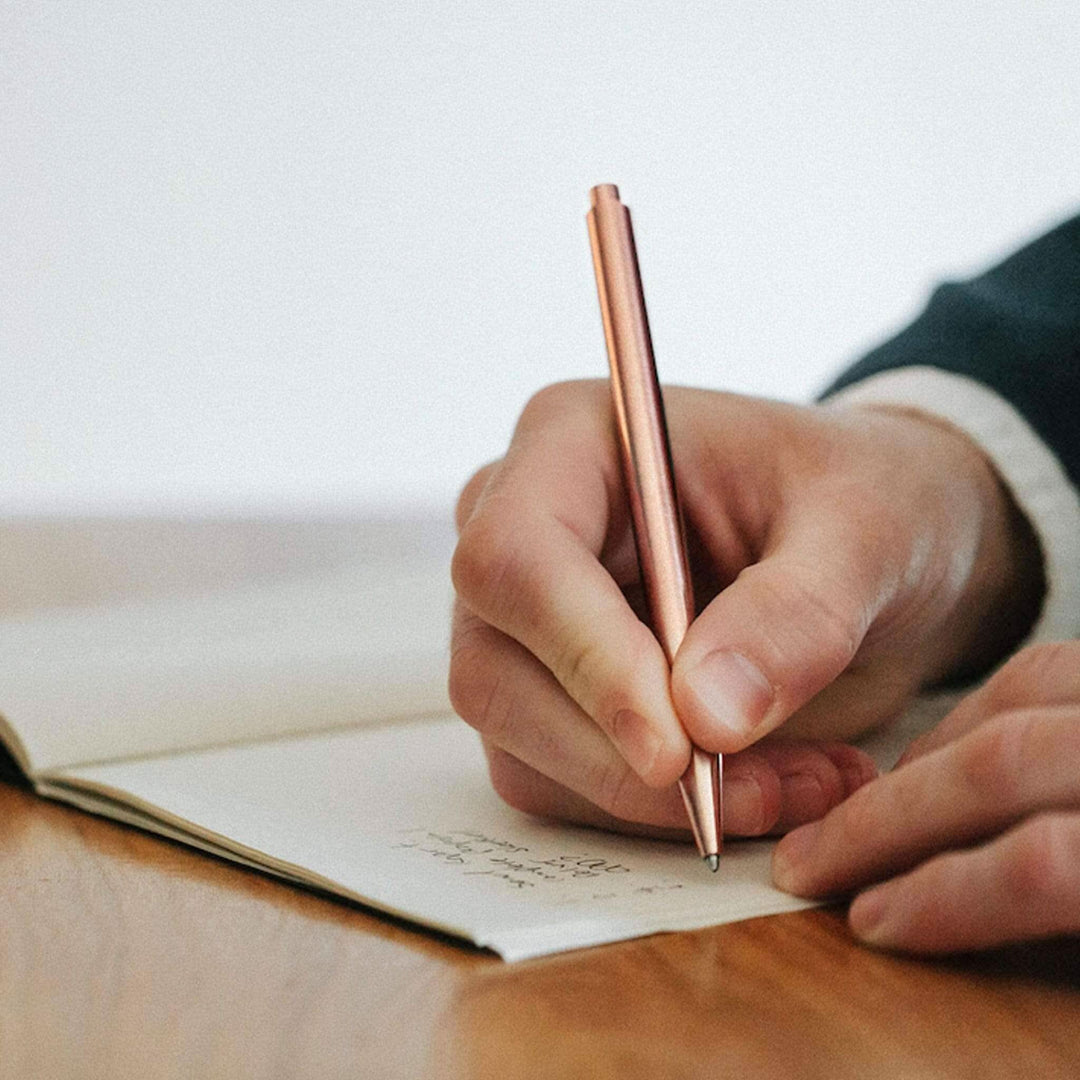 Stylo Modern Fuel en métal rose, utilisé pour écrire dans un carnet sur une table en bois.