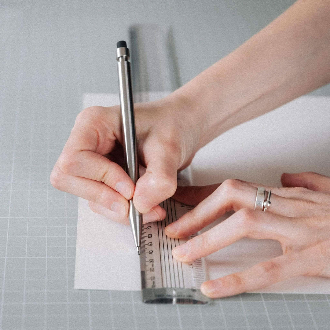 Person using a silver ruler and pen to draw on a sheet of paper in a creative workspace.