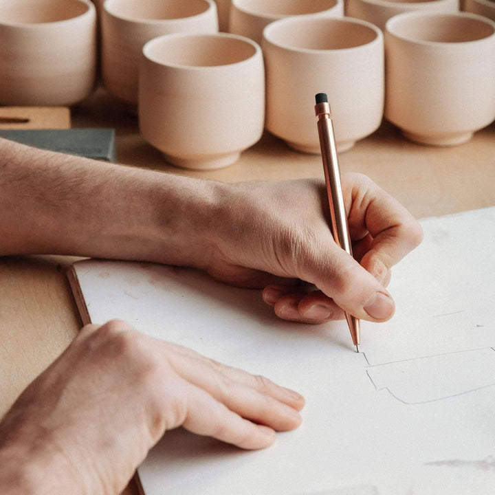 Un artisan dessinant avec un crayon Modern Fuel sur une feuille, des pots en argile flous en arrière-plan.