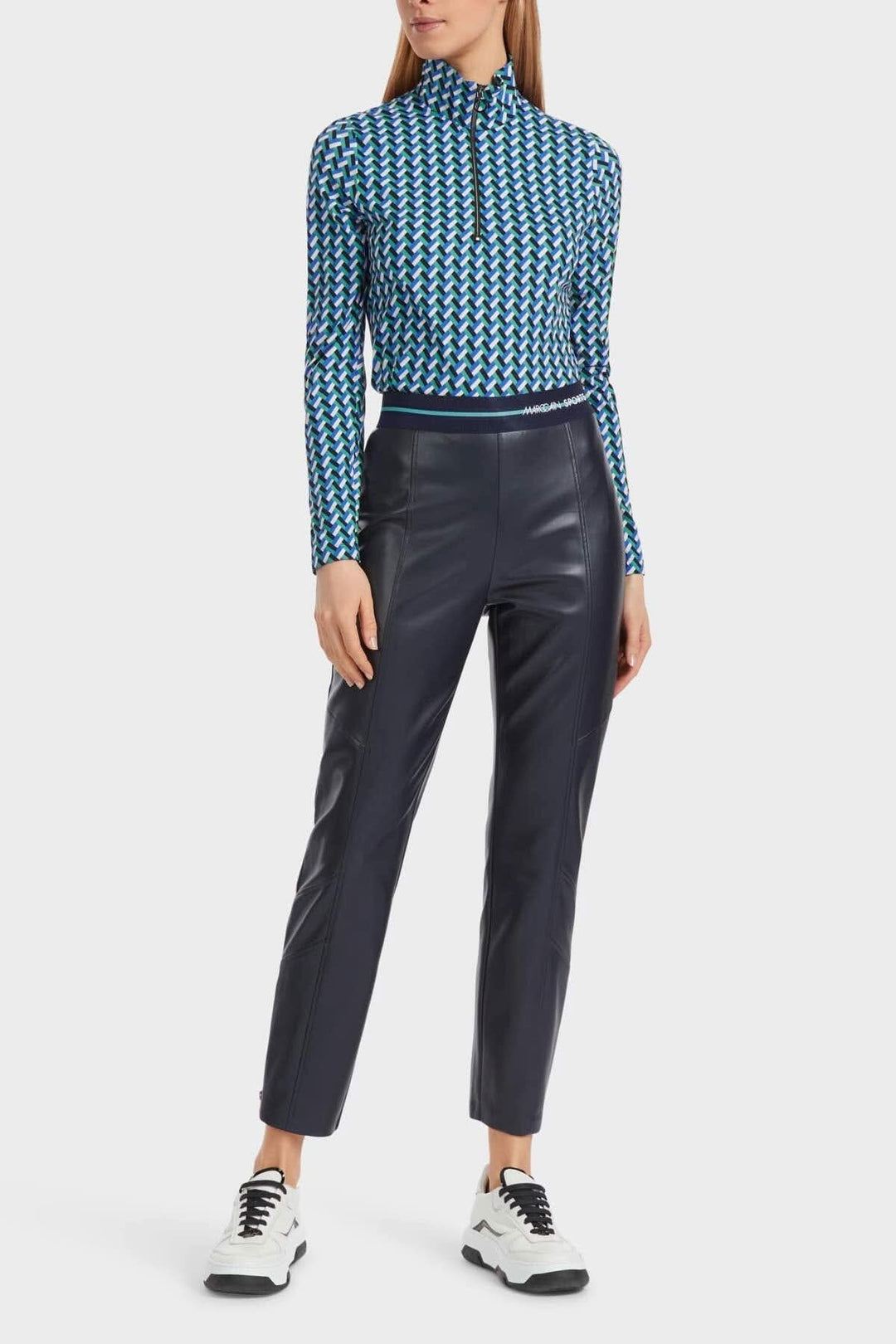 Femme en pantalon en cuir noir et haut à motifs géométriques, chaussée de baskets blanches.
