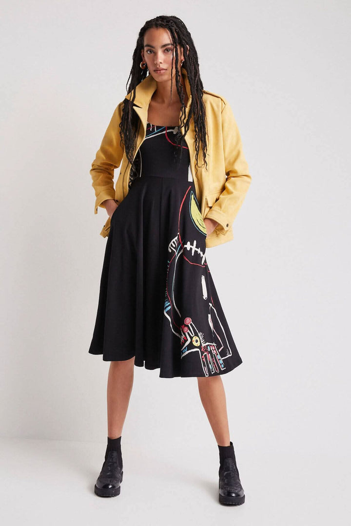 Femme en robe noire midi à imprimé artistique portant une veste en cuir jaune, pose élégante sur fond gris.
