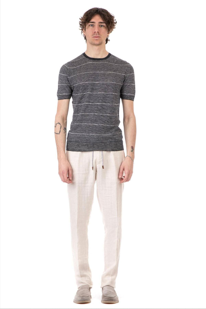 Homme portant un t-shirt à rayures noires et grises avec col rond, accompagné de pantalons légers blancs.