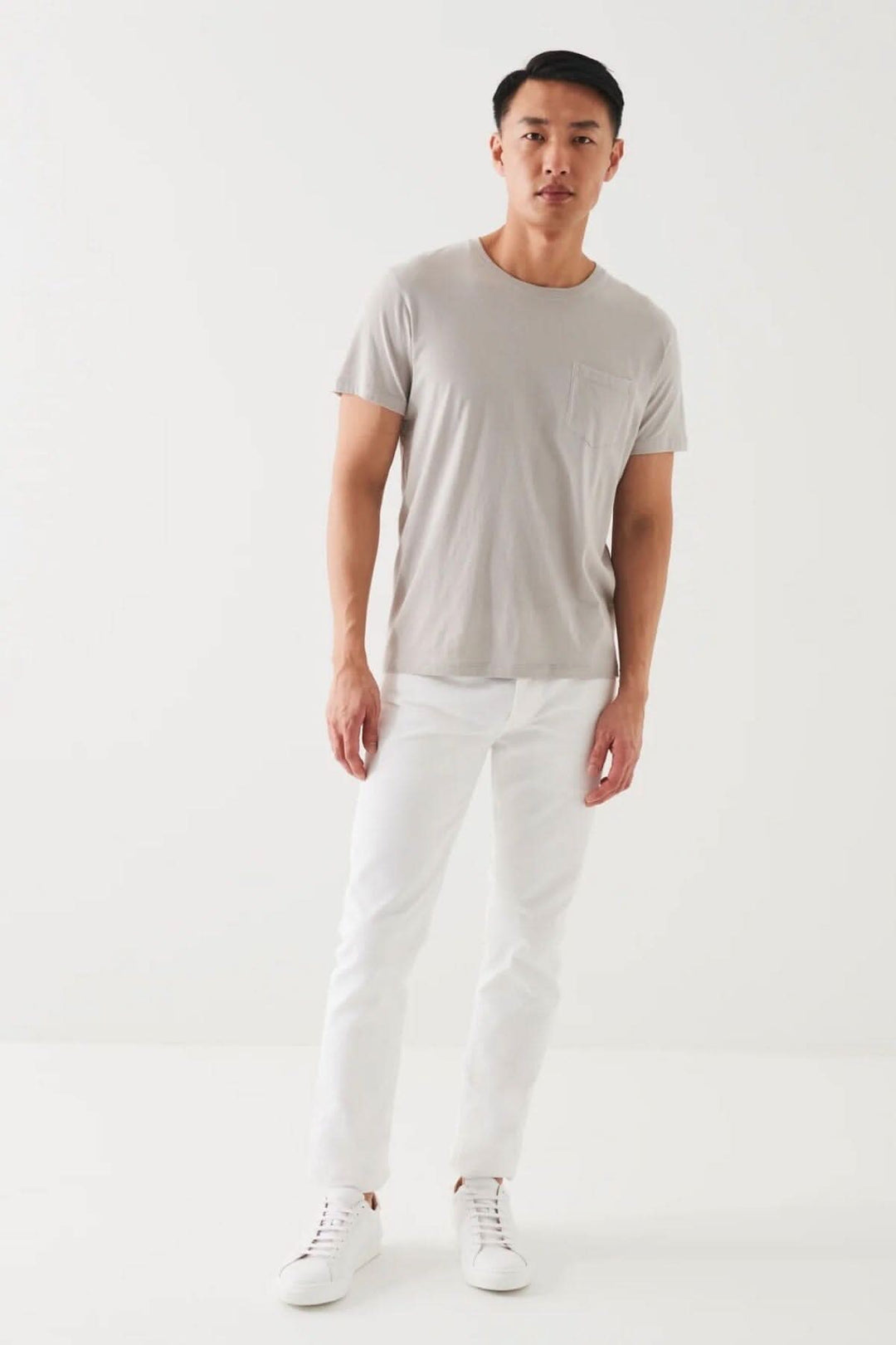 Homme portant un t-shirt léger en coton pima gris avec une poche, accompagné de jeans blancs et de chaussures blanches.